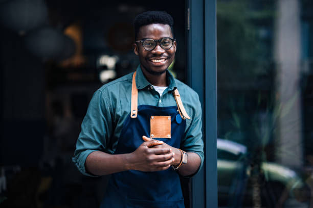 Cheerful smiling African-American barista wearing an apron and standing at the entrance of his cafe and looking at camera.