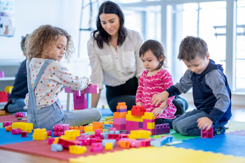 A young female childcare worker sits on the floor as she plays with the children.  They have scattered blocks on the floor that they are using together to build with.